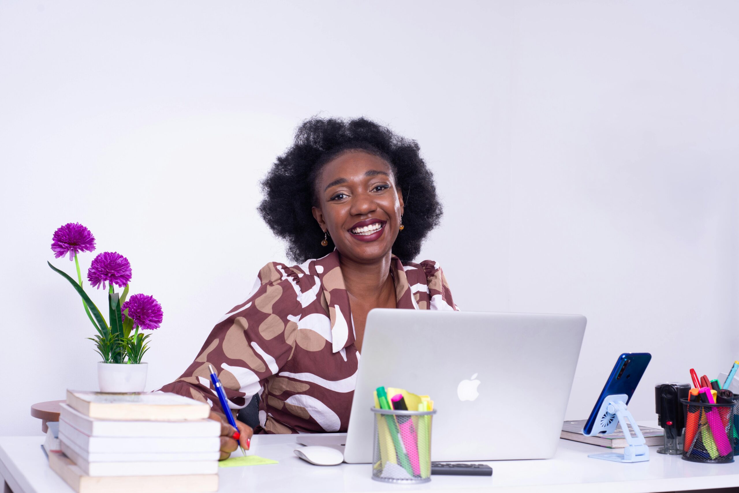 A young female Nigerian finance professional with an Afro sits at a desk with her laptop, smiling and with the best finance books of 2025 stacked on her desk beside her.