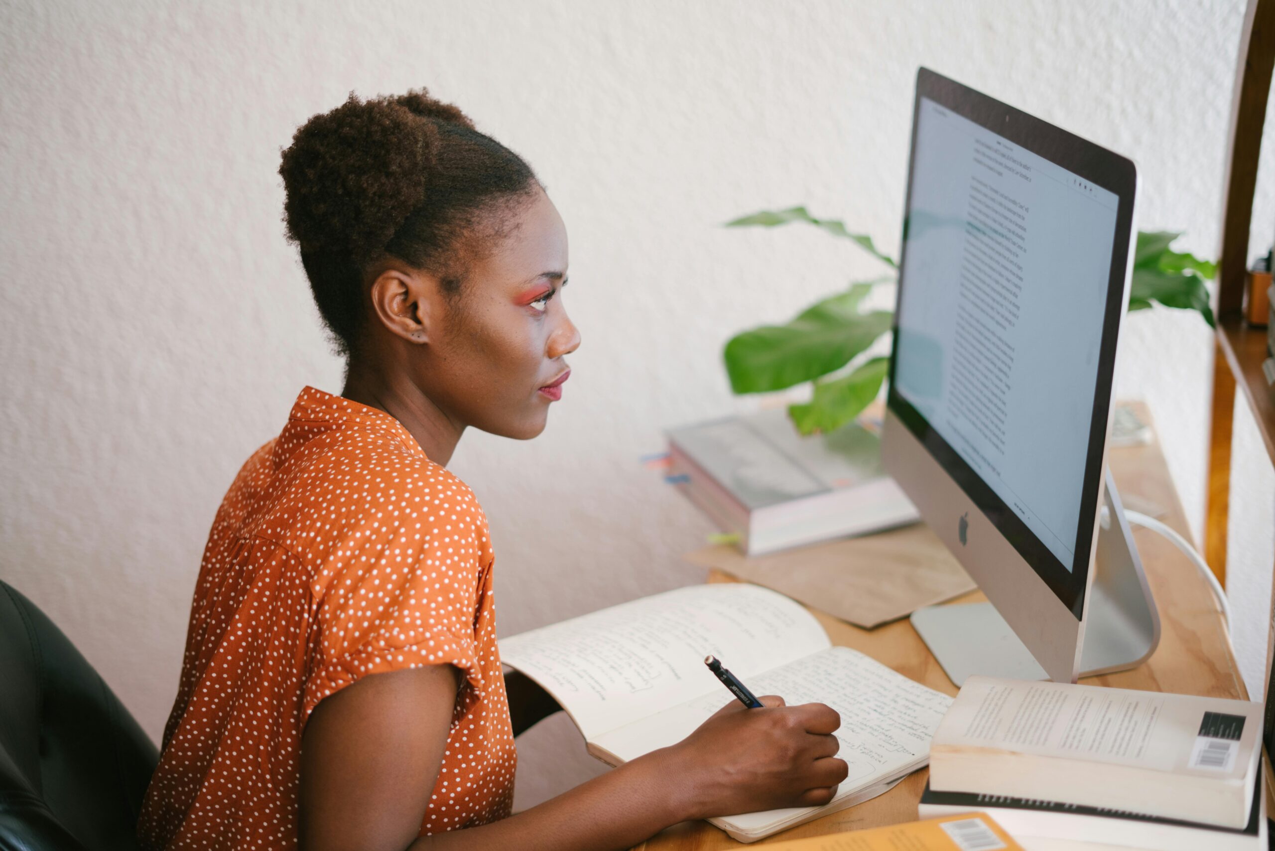 A focused finance professional sits at a desk, writing notes in a journal while working on a desktop computer. She is surrounded by books, reflecting a dedication to learning about finance courses and certifications, possibly exploring 2025 finance courses or certifications for Nigerian accountants.